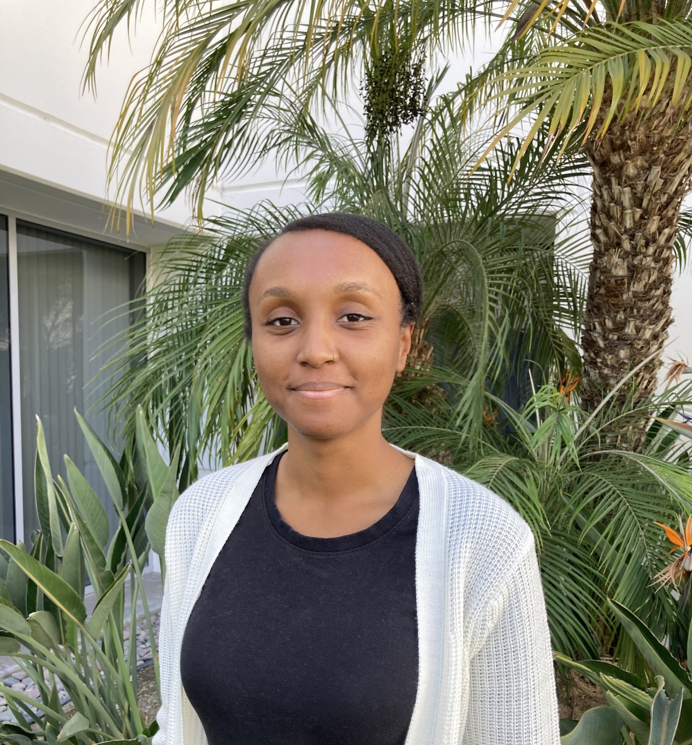 African American, Black female standing in front of green plant in front of white building. She has on a white cotton sweater over a black shirt.